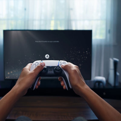 Pessoa segurando um controle sem fio DualSense enquanto configura um console PlayStation 5, com a tela da TV exibindo a mensagem ‘Pressione o botão PS no seu controle.