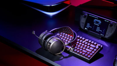 Audeze Maxwell 2 headset on a gaming desk next to a keyboard and handheld device with blue and red lighting.
