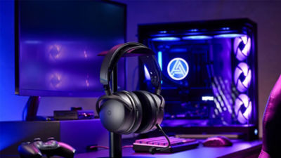 Close-up of Audeze Maxwell 2 headset on a stand in a desktop setup with keyboard and LED-lit PC tower.