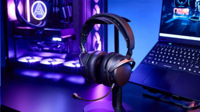 Audeze Maxwell 2 headset on a stand next to a laptop and LED gaming PC with blue lighting.