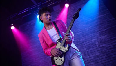 Player wearing a pink jacket performing with a white Riffmaster wireless guitar controller under stage lights.