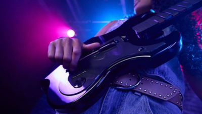 Close-up of a player’s hand strumming the black Riffmaster guitar controller under blue and pink lights.